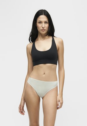 Femme aux longs cheveux foncés portant un soutien-gorge de sport noir et un sous-vêtement taille haute beige clair, debout devant un fond clair uni.