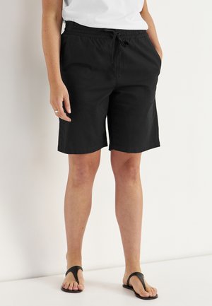 Person iført sorte knælange shorts med snøre i taljen, hvid top og sorte sandaler, stående op ad en hvid væg.