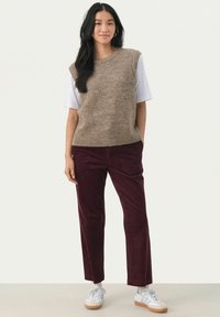 Mouwloze beige gebreide trui over een wit shirt, gecombineerd met burgundy corduroy broek en witte sneakers. Heeft ribbenrand. 