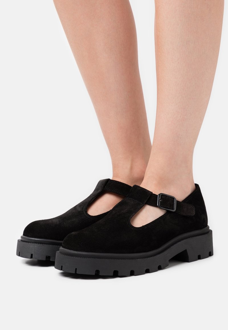 Chaussures à brides en T en suède noir avec semelles épaisses à crampons et boucle latérale réglable, portées pieds nus sur un fond clair uni.