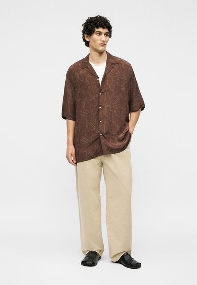 Άνδρας όρθιος, φοράει καφέ πουκάμισο με κοντά μανίκια και μοτίβο, μπεζ παντελόνι με φαρδιά μπατζάκια, μαύρα σανδάλια και λευκό φανελάκι, κοιτάζει πλευρικά.