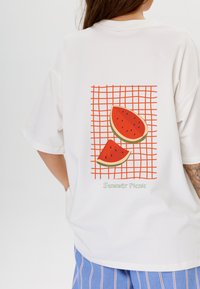 Witte t-shirt met een grafisch ontwerp van watermeloenplakken op een rode raster achtergrond, met de tekst "Zomer Picknick" in het groen onder het ontwerp.