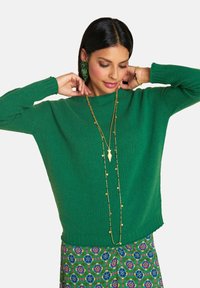 Frau, die eine lange goldene Halskette anpasst, trägt einen grünen Pullover, einen blumigen Rock und passende grüne Perlenohrringe vor einem weißen Hintergrund.