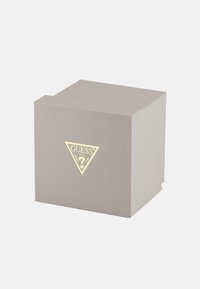 Siv teksturiran kvadratni škatla z zlatim logotipom Guess, ki na sprednji strani prikazuje trikotnik in vprašaj.