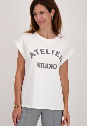Vrouw met bruin haar in een witte shirt met opgerolde mouwen met de tekst "ATELIER STUDIO" en grijze broek, glimlachend en kijkend naar de zijkant.