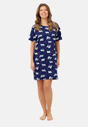 Uśmiechnięta kobieta stojąca boso, ubrana w granatową sukienkę z białymi wzorami kotów i różową piłeczką z włóczki na jednolitym tle.