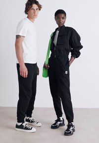 Twee modellen staan naast elkaar. Het mannelijke model draagt een wit T-shirt en zwarte joggingbroek. Het vrouwelijke model draagt een zwarte ritsjack en bijpassende joggers, terwijl ze een felgroene tas vasthoudt. De schoenen zijn zwart en wit.