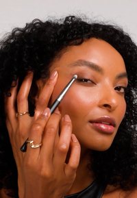 Make-upkwast met een metalen zilverkleurig handvat, zachte haren; model brengt make-up aan, draagt gouden ringen aan de vingers, zwarte top. Soepele huid, natuurlijke glans.