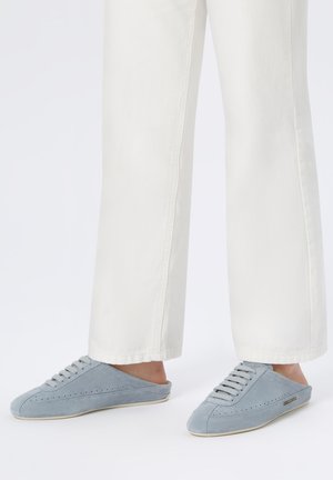 Persona con pantaloni bianchi e sneaker slip-on azzurre con lacci, in piedi su una superficie bianca.