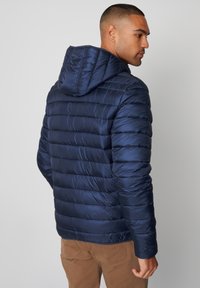Giacca puffer blu navy con cappuccio, caratterizzata da baffi orizzontali, una texture lucida e polsini elasticizzati. Vestibilità casual adatta per essere indossata a strati.