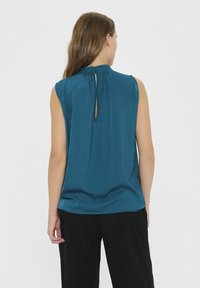 Top senza maniche color teal con dettaglio a oblò con bottone sulla schiena. Tessuto morbido con un fit rilassato. Abbinato a pantaloni neri, design minimalista.