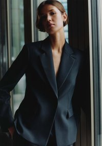 Blazer nero su misura con rever affilati, spalle strutturate e scollatura a V profonda. Tessuto liscio con un leggero lucido, silhouette aderente.