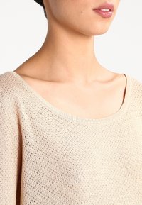 Ljust beige stickad tröja med lös passform och vid rund halsringning. Tyget har ett strukturerat öppet vävmönster.