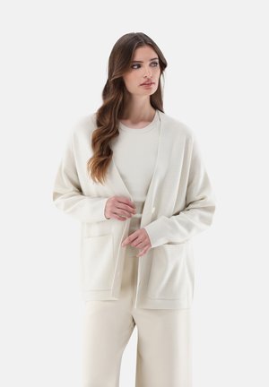 Hellbeige Strickjacke mit weicher Textur, offenen Vorderseite, zwei aufgesetzten Taschen und großen Knöpfen, getragen über einem passenden Top und einer Hose.