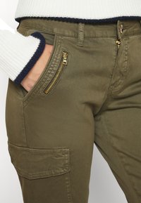 Calças cargo verde-azeitona com uma textura suave, apresentando fechos de correr dourados e um bolso lateral. Detalhe de costura perto da cintura.