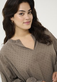 Lichtbruine blouse van een lichtgewicht materiaal met een v-hals en rimpelaccenten, versierd met gestructureerde stippenpatronen. Lange mouwen met aangesloten manchetten. Soepele stof.
