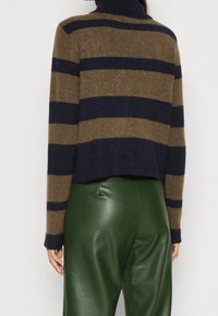 Vrouw in een bruin en marineblauw gestreept wijde trui en hoogopgetrokken donkergroene leren broek, gezien vanuit de rug.