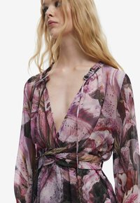 Langärmliges Kleid mit tiefem V-Ausschnitt, floralem Print in Pink und Fuchsia sowie einer gebundenen Taillenpartie, aus leichtem, durchscheinendem Stoff.