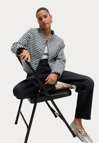 Giacca houndstooth in bianco e nero con colletto, abbinata a jeans neri a vita alta e ballerine metalliche argento, seduta su una sedia nera.