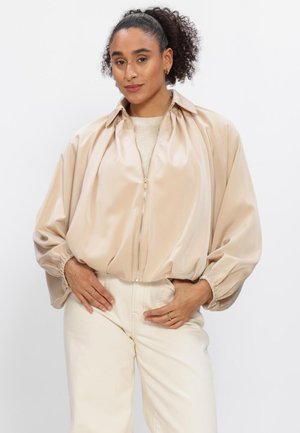 Femme aux cheveux bouclés portant une veste beige à manches ballon zippée et un pantalon blanc cassé, debout les mains sur les hanches sur fond blanc.