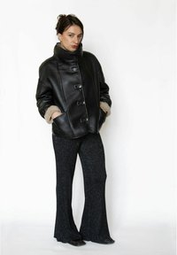 Sort læderjakke med høj krave, der har en knaplukning foran og pelsforing ved manchetterne. Kombineret med glitrende sorte udvidede bukser.