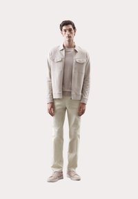 Hellbeige Zip-Jacke mit zwei Brusttaschen, übereinander getragen mit einem passenden Rundhalsausschnitt-Shirt, kombiniert mit hellen, weißen Hosen und hellen Sneakern.