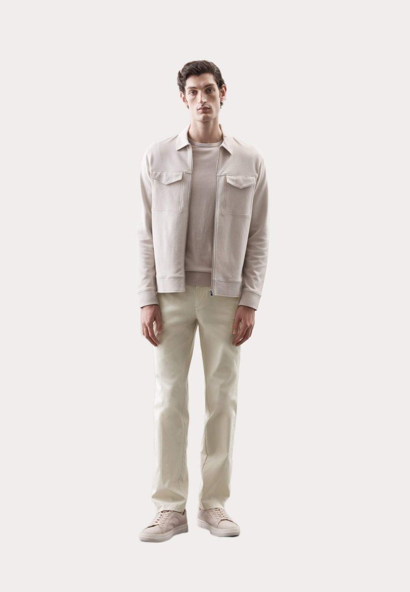 Hellbeige Zip-Jacke mit zwei Brusttaschen, übereinander getragen mit einem passenden Rundhalsausschnitt-Shirt, kombiniert mit hellen, weißen Hosen und hellen Sneakern.