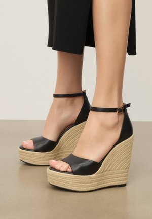 Feet wearing black leather ankle-strap wedge sandals with open toes and woven jute platform soles, paired with black cropped pants.