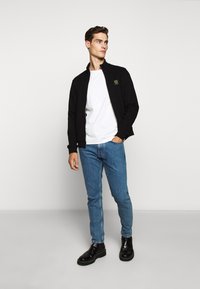 Svart zip-jacka med ribbade manschetter, bärs över en vit T-shirt. Modellen matchar den med ljusblå jeans och svarta läderstövlar.