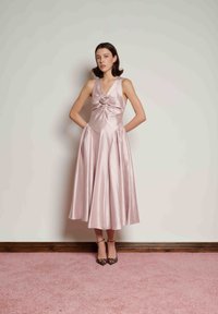 Γυναίκα με αμάνικο midi φόρεμα από απαλό ροζ σατέν με λουλουδένια λεπτομέρεια στο στήθος, που στέκεται σε ροζ χαλί μπροστά σε λευκό τοίχο, φορώντας τακούνια με λεοπάρ.