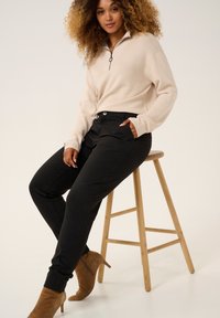 Béžový pulóver na zips s ribovanou textúrou v štvrť dĺžke, skombinovaný s čiernymi nohavicami so bočnými vreckami. Outfit dopĺňajú hnedé nubukové členkové topánky.