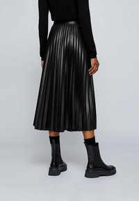 Gonna midi plissettata nera realizzata in materiale sintetico, abbinata a stivaletti neri chunky e calze nere a coste, vista da dietro.