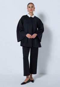 Črna jakna z širokimi rokavi in belim ovratnikom, v kombinaciji s krojenimi črnimi hlačami in črnimi pointed flats čevlji. Gladek material z minimalnimi detajli.