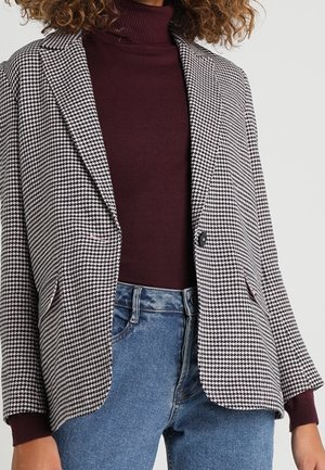 Femme portant un blazer pied-de-poule noir et blanc sur un pull à col roulé bordeaux et un jean en denim bleu.