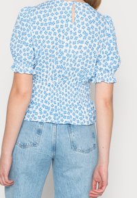 Haut blanc à motif floral bleu, taille smockée, manches bouffantes et découpe en forme de clé à l'arrière ; associé à un jean taille haute bleu clair.