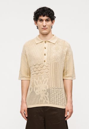 Mladý muž s kučeravými vlasmi v béžovej pletenej polokošeli s krátkym rukávom s patchworkovým vzorom a tmavohnedými nohavicami, stojaci.