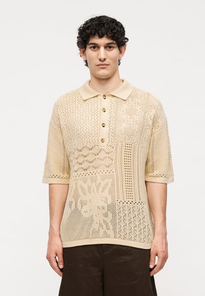 Jeune homme aux cheveux bouclés portant un polo en maille beige à manches courtes avec un motif patchwork et un pantalon marron foncé, debout.