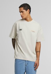 Beige t-skjorte med korte ermer, med svart broderi "out of office" og oransje skriftakenter. Enkelt design og avslappet passform.