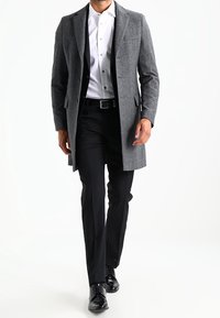 Hombre caminando vestido con un abrigo gris, una camisa blanca, pantalones negros, cinturón negro y zapatos de vestir negros pulidos, contra un fondo blanco.