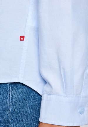 Gaiši zilas krāsas garām piedurknēm krekla aproce ar pogu, sarkans tagiņš ar baltu zvaigzni blakus sānu šuvēm, valkāts virs zilām džinsa biksēm.