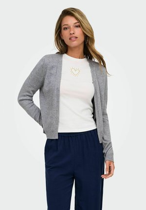 Kvinde med løst blond hår iført grå åben cardigan, hvid T-shirt med gyldent hjerte-design og marineblå bukser, står mod hvid baggrund.