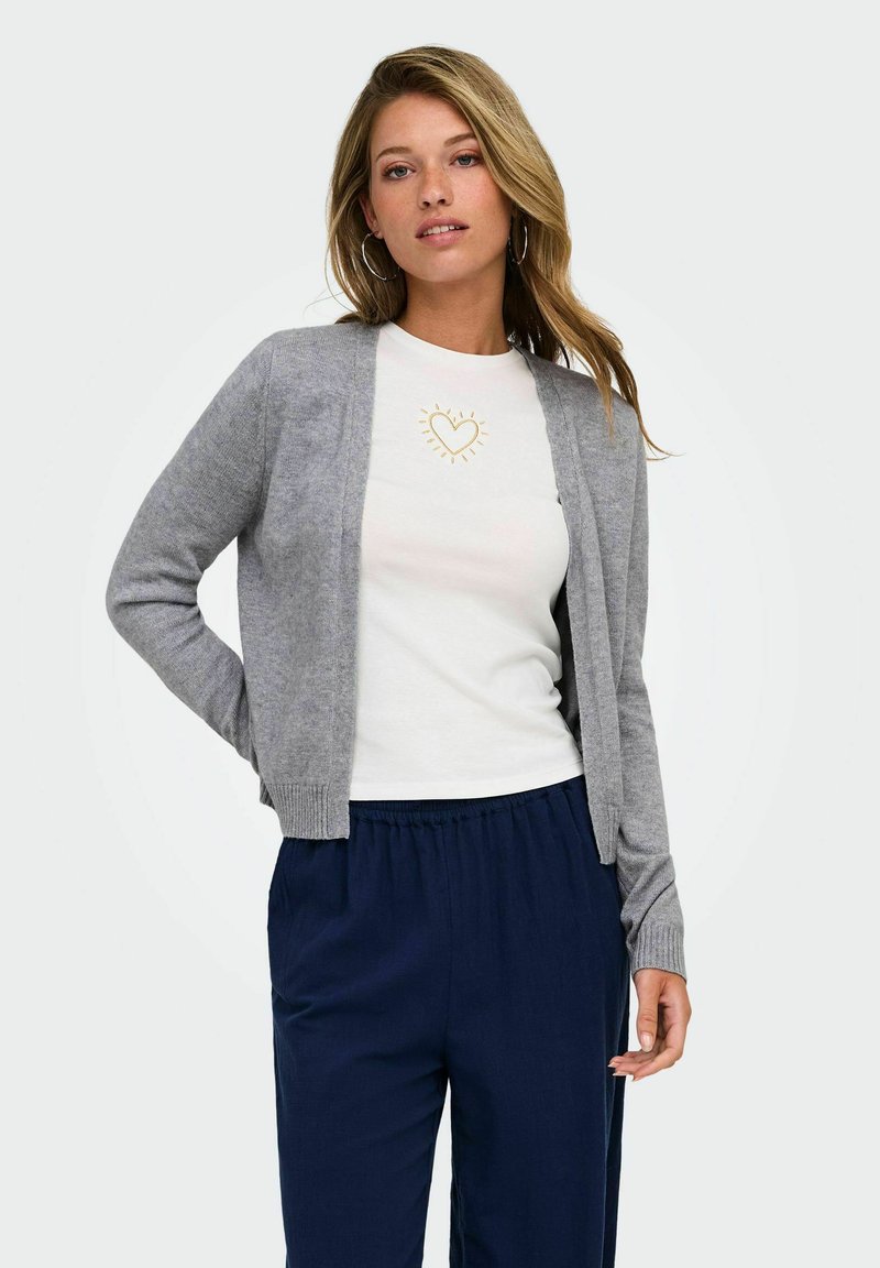Femme aux cheveux blonds lâchés porte un cardigan gris ouvert, un T-shirt blanc avec un motif cœur doré et un pantalon bleu marine, debout sur fond blanc.