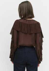 Ruskea sifonkipaita, jossa on off-shoulder-tyylinen röyhelödesign, pitkät hihat, joissa on röyhelöitä hihansuissa, yhdistettynä tummiin farkkuihin.