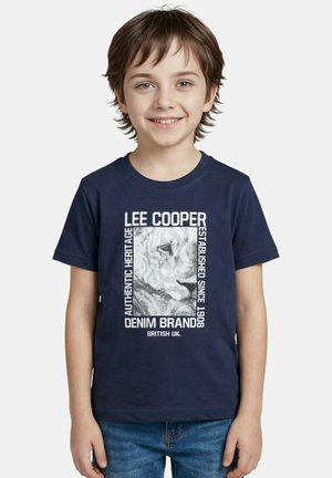 Garçon souriant aux cheveux bruns portant un t-shirt bleu marine avec un texte blanc et un motif, assorti à un jean bleu.