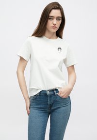 Biele bavlnené tričko s krátkymi rukávmi, na ľavej strane na hrudi s malým grafickým motívom osoby s kučeravými vlasmi. Kombinované s modrými džínsami.