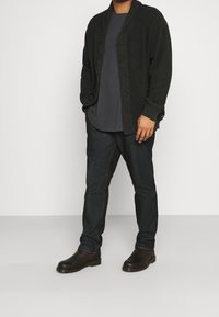 Dunkelgrüner Cardigan mit gerippter Struktur, Knopfverschluss, über einem grauen T-Shirt getragen, kombiniert mit dunkelblauen Jeans und braunen Stiefeln.