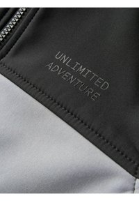 Schwarzer und grauer Stoff mit glatter Textur; enthält den text "UNLIMITED ADVENTURE" in grauem Faden in der Nähe des Reißverschlusses gestickt.