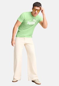 T-shirt verde in cotone con logo bianco Puma, abbinata a pantaloni larghi color crema e sneaker beige; modello che indossa occhiali da sole con lenti gialle.