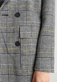 Grå rutigt kavaj med en texturerad tyg och svarta och gula detaljer, dubbelknäppt design och en enda bröstficka.