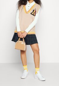 Beige stickad väst med gul och orange kant; ett märke; vit långärmad skjorta; rutig kjol; beige handväska; gula randiga strumpor; vita sneakers.